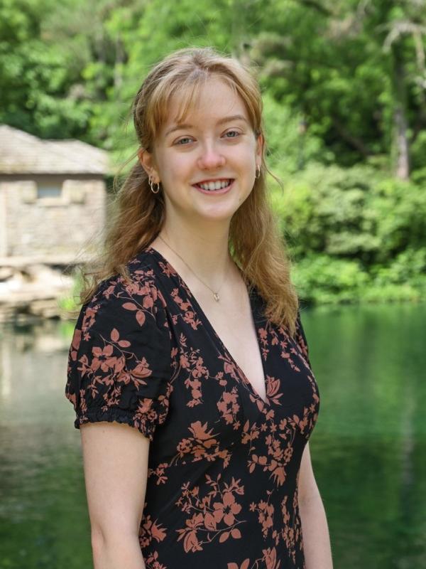 Sophie Papp, a white person with chest length blonde hair, standing in front of a pond and smiling.